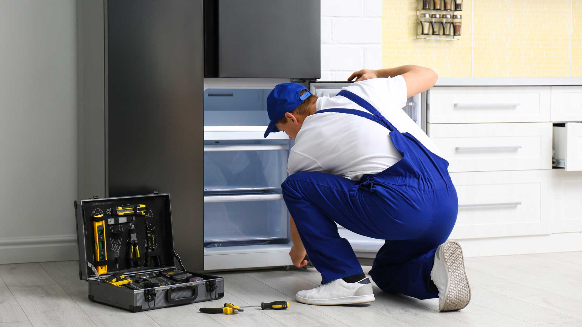 A man in blue overalls kneeling to inspect a refrigerator