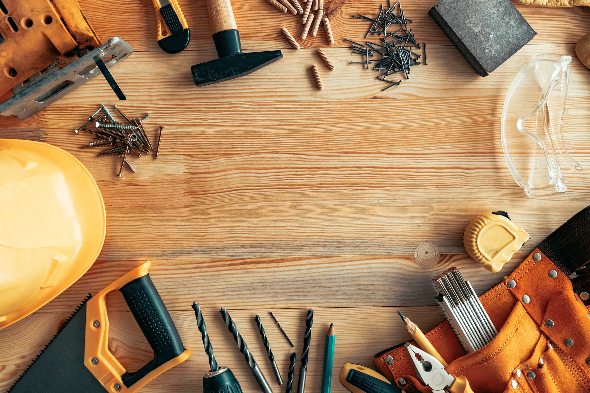 Tools scattered over a bench, including a hardhat, safety goggles, drill bits, nails, screws, a handsaw, a hammer, a sanding block, and a tape measure