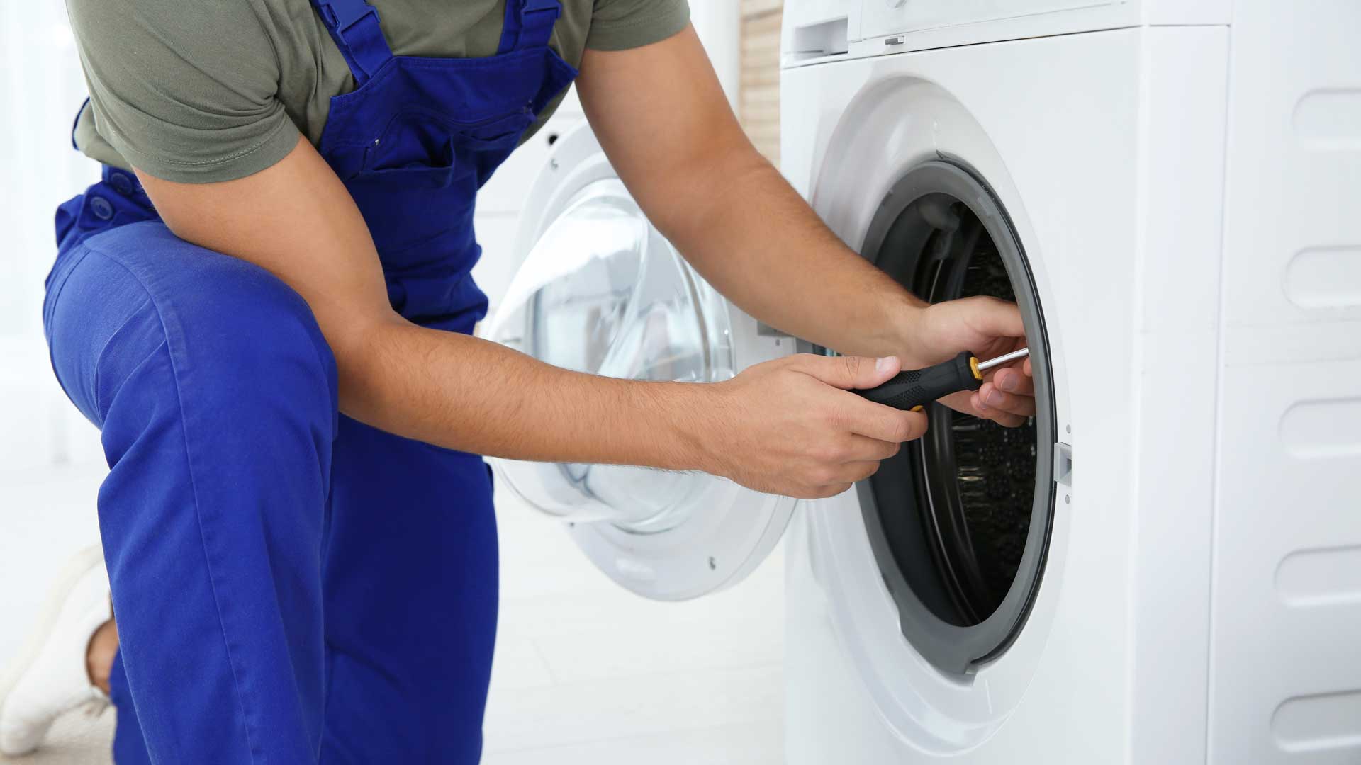 A repair man in blue overalls fixing a washer/dryer